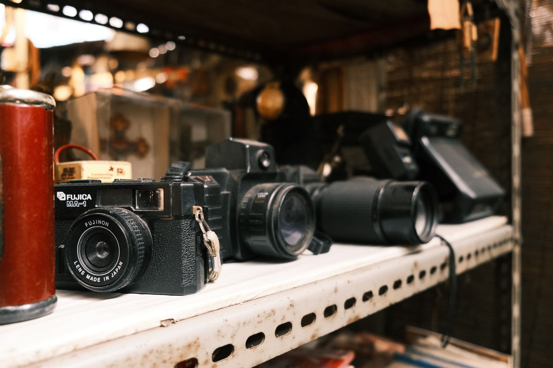 Appareils photo argentiques anciens, dont un Fujica MA-1, alignés sur une étagère métallique blanche usée dans un stand d’objets d’occasion.