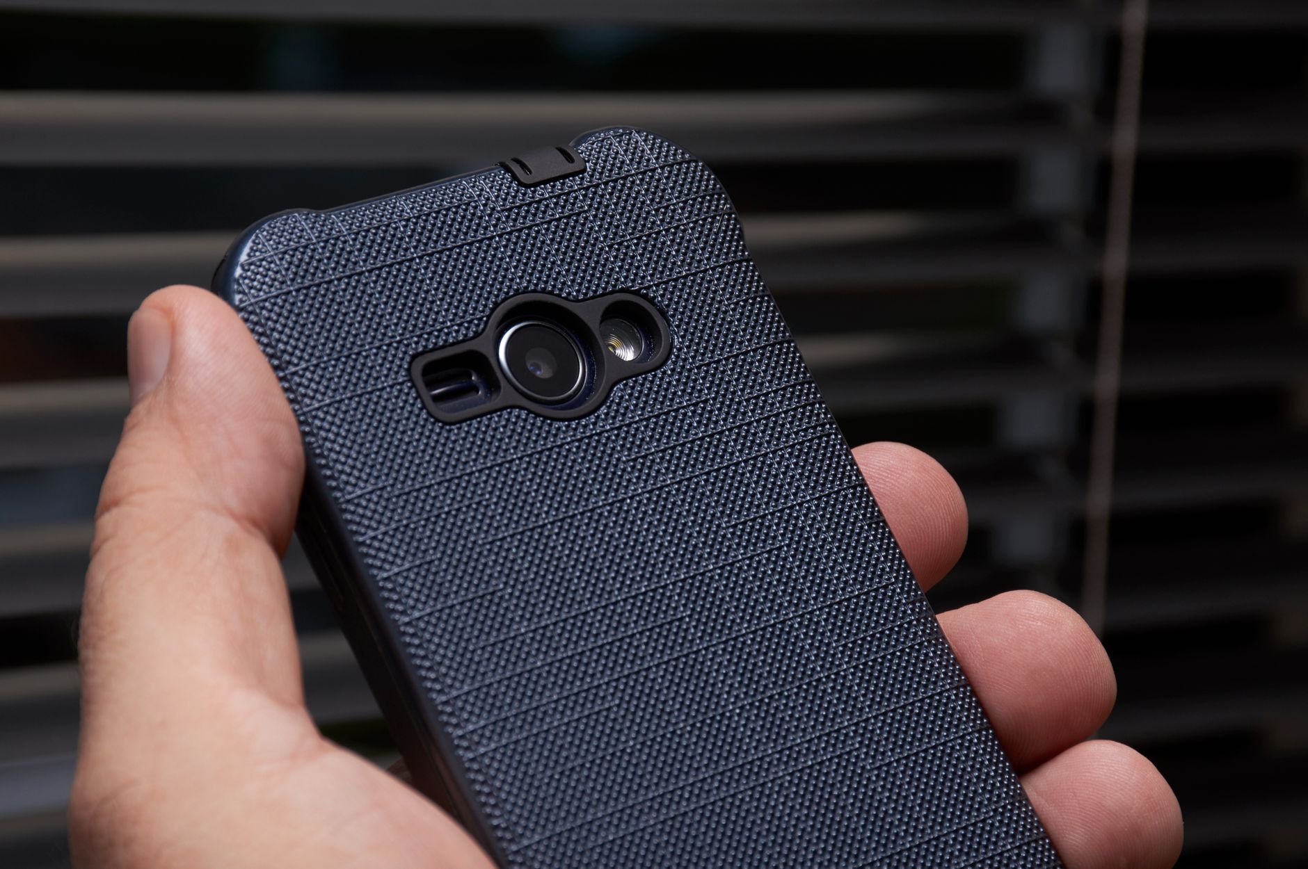 Main tenant un smartphone dans une coque texturée bleu foncé, vu de dos, avec un petit objectif et un flash.