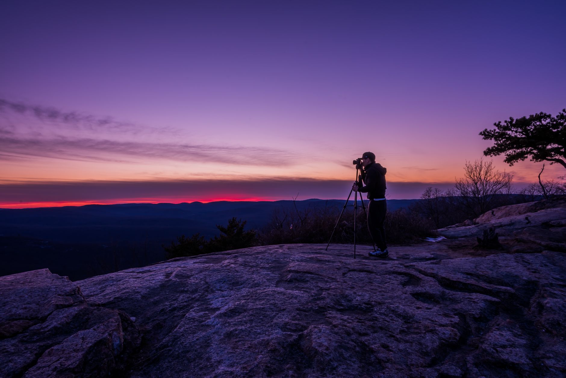 Photographe en silhouette sur un rocher, appareil monté sur trépied, face à des collines sous un ciel de crépuscule violet et rouge.