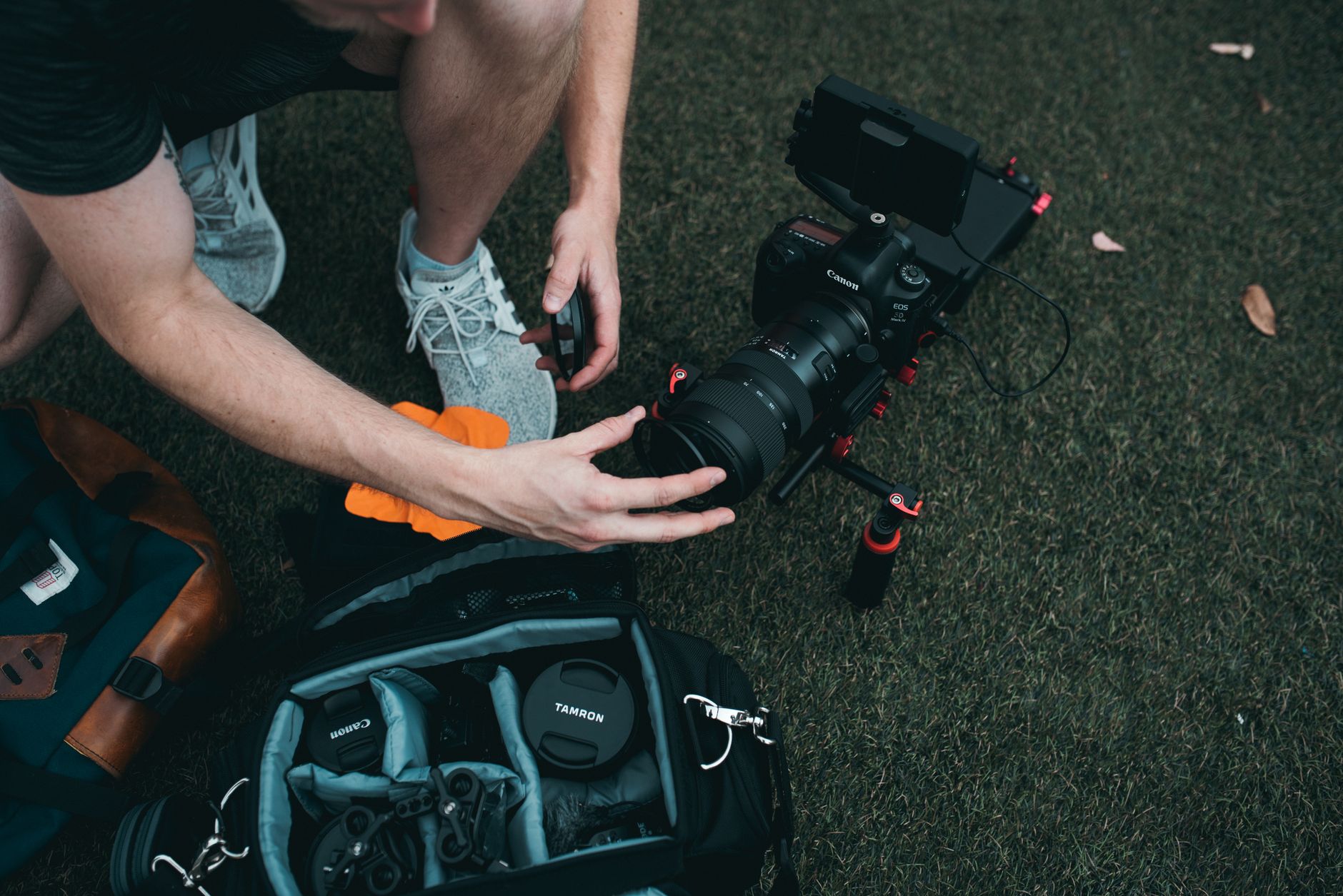 Un homme accroupi sur l’herbe manipule l’objectif d’un appareil photo Canon sur cage, à côté d’un sac photo ouvert contenant du matériel.